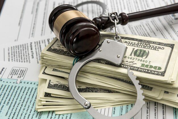 A judge's gavel and handcuffs rest on a stack of hundred-dollar bills and tax forms, symbolizing financial crime.